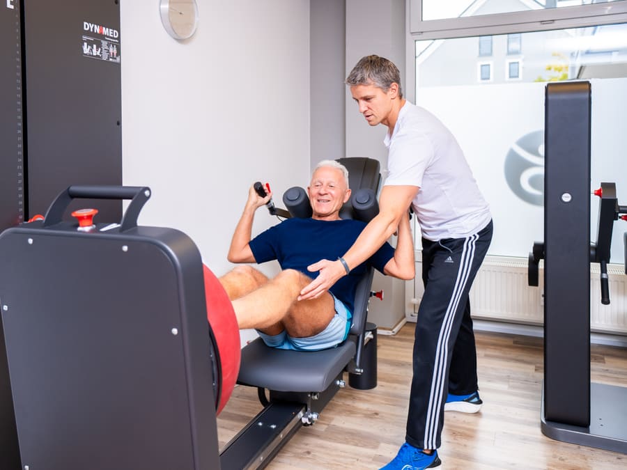 Patient trainiert an einem großen KGG-Gerät (Beinpresse), ein Therapeut korrigiert die Übung